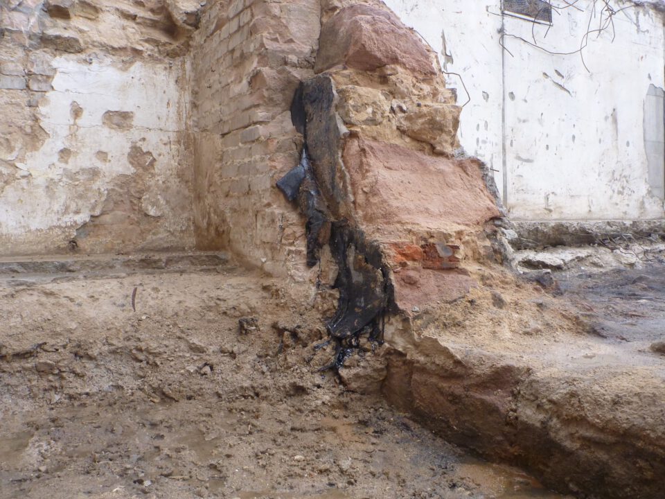 Das Bild zeigt eine freigelegte Mauer aus grob behauenen Sandsteinblöcken und Ziegeln, die teilweise beschädigt ist. An der Wand ist eine dunkle, teerartige Abdichtung zu erkennen, die in Streifen von oben nach unten verläuft. Der Boden davor besteht aus feuchtem Erdreich und wirkt uneben, während die Hintergrundwand stark verwittert und fleckig ist.