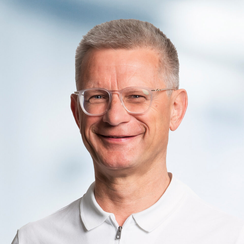 Das Bild zeigt ein professionelles Porträt eines Mannes mit sehr kurzem grauem Haar und einer hellen, transparenten Brille. Er lächelt freundlich in die Kamera und trägt ein weißes Poloshirt mit Reißverschluss am Kragen. Der Hintergrund ist weich verlaufend in hellen Blau- und Weißtönen gehalten und wirkt ruhig und neutral.