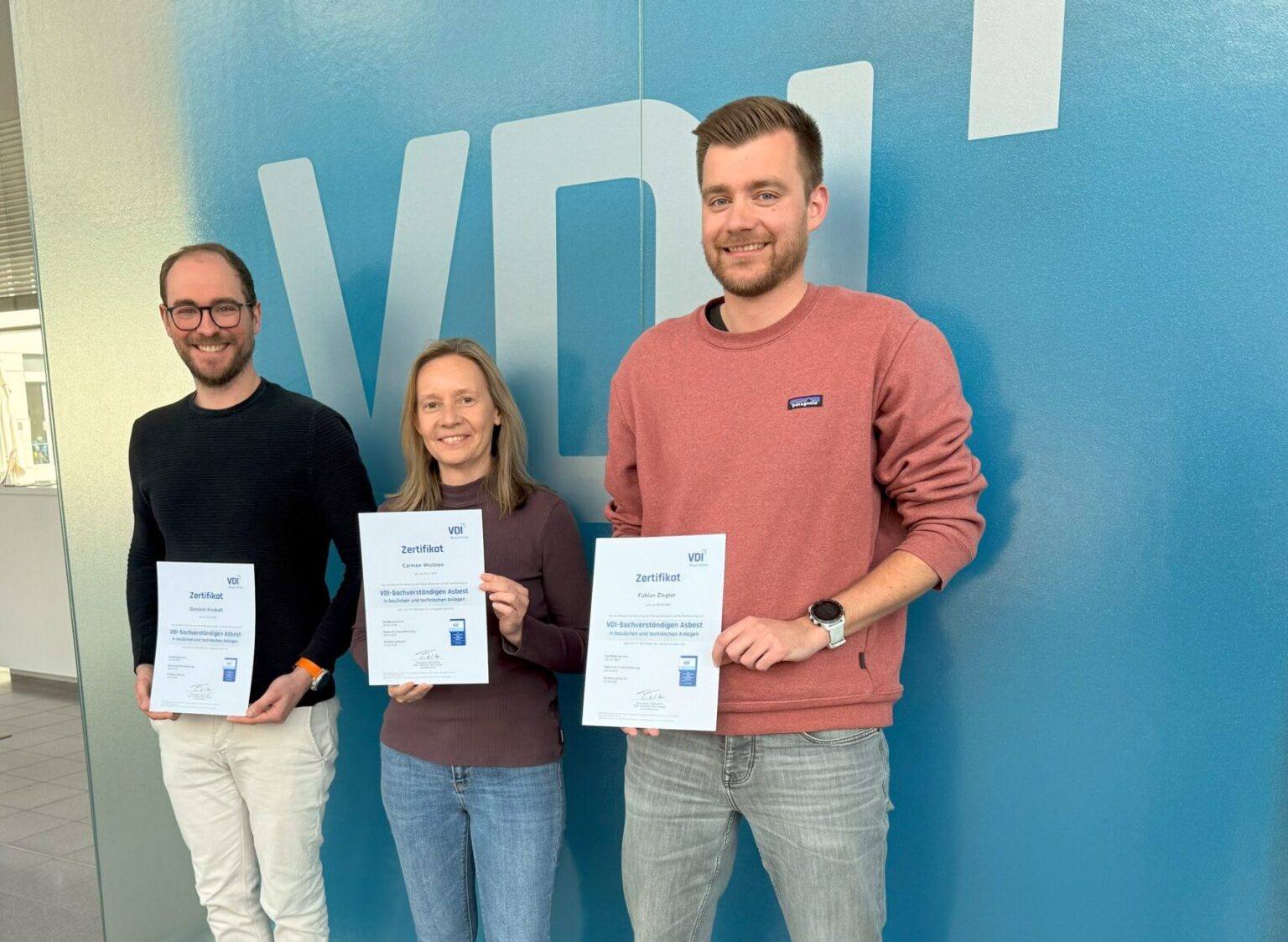 Das Bild zeigt drei Personen, die nebeneinander vor einer großen blauen Wand mit einem großen VDI-Logo stehen. Jede Person hält ein Zertifikat in den Händen, das auf eine erfolgreiche Qualifikation hinweist. Die Kleidung ist leger, und die Szene wirkt wie eine offizielle Übergabe oder ein Fototermin zur Zertifizierung.