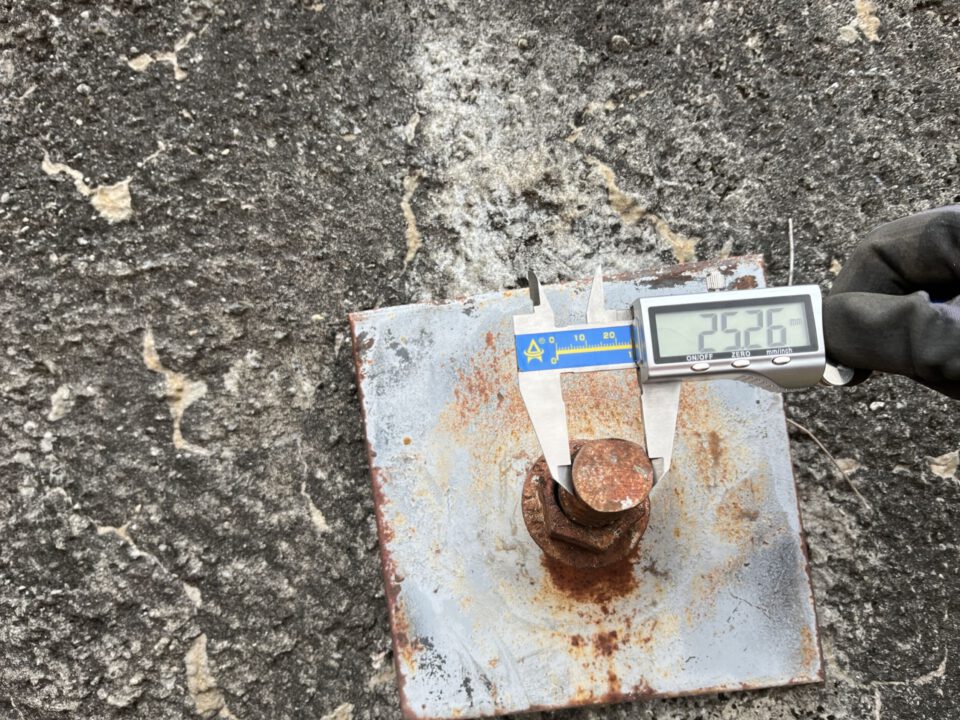 Das Bild zeigt eine Nahaufnahme einer rostigen Metallplatte mit einem großen Bolzen, der in eine Felsoberfläche eingelassen ist. Auf dem Bolzen wird mit einer digitalen Schieblehre der Durchmesser gemessen, wobei das Display den Wert anzeigt. Die Oberfläche des Felsens ist grau und rau, und die Metallteile weisen deutliche Rostspuren auf.