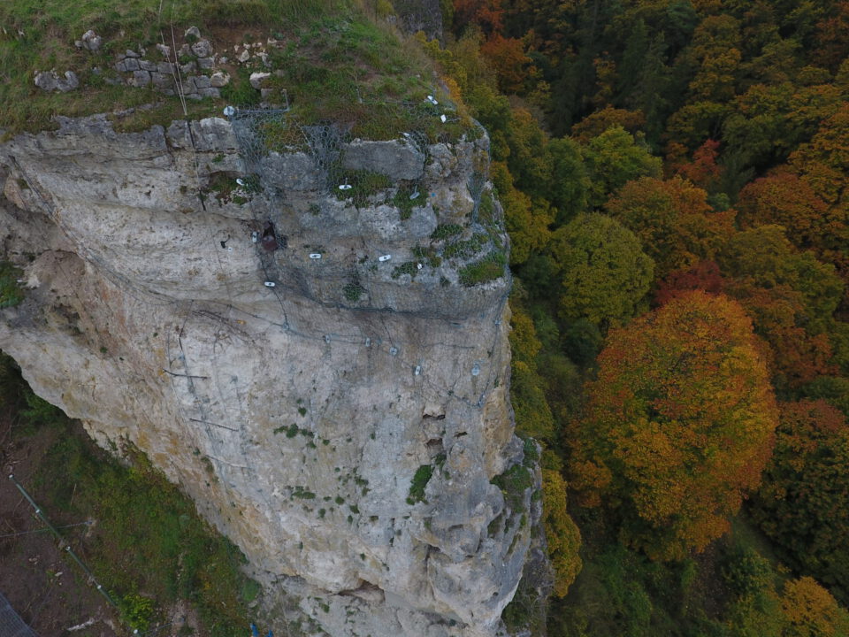 Das Bild zeigt eine steile Felswand, die von oben bis unten mit einem Drahtnetz gesichert ist. Auf der Felsoberfläche sind mehrere Befestigungspunkte sichtbar, die das Netz fixieren, und am oberen Rand wächst Gras und kleine Vegetation. Im Hintergrund erstreckt sich ein dichter Wald mit Bäumen in herbstlichen Farben wie Grün, Gelb und Orange.