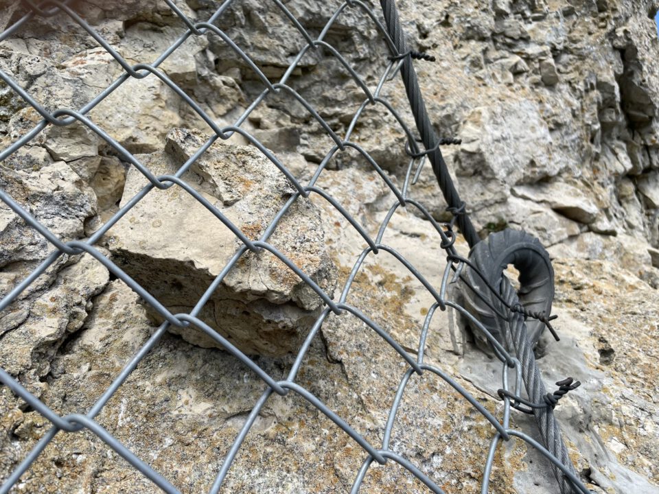 Das Bild zeigt eine Felswand, die mit einem engmaschigen Drahtnetz gesichert ist. Hinter dem Netz liegt ein kantiger Stein, der von den Metallmaschen zurückgehalten wird. Rechts ist ein dickes Stahlseil zu sehen, das durch eine runde Metallöse geführt und mit weiteren Seilen befestigt ist, um die Konstruktion zu stabilisieren.