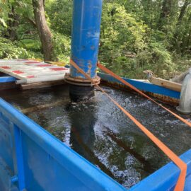 Das Bild zeigt einen großen blauen Wasserbehälter, der mit Wasser gefüllt ist und in dem sich deutliche Strömungen an der Oberfläche bilden. Ein dickes Metallrohr führt von außen in den Behälter und leitet Wasser hinein, während ein senkrechtes blaues Rohr in der Mitte befestigt ist und mit orangefarbenen Gurten gesichert wird. Im Hintergrund sind grüne Bäume und Sträucher zu sehen, was auf eine Installation im Freien hinweist.