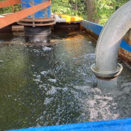 Das Bild zeigt einen großen blauen Wasserbehälter, der mit Wasser gefüllt ist und in dem sich deutliche Strömungen und Blasen an der Oberfläche bilden. Rechts mündet ein dickes Metallrohr in den Behälter und leitet Wasser hinein, während im Hintergrund ein weiteres Rohr und orangefarbene Gurte zur Befestigung sichtbar sind. Die Szene befindet sich im Freien, umgeben von grünem Laub und Bäumen.