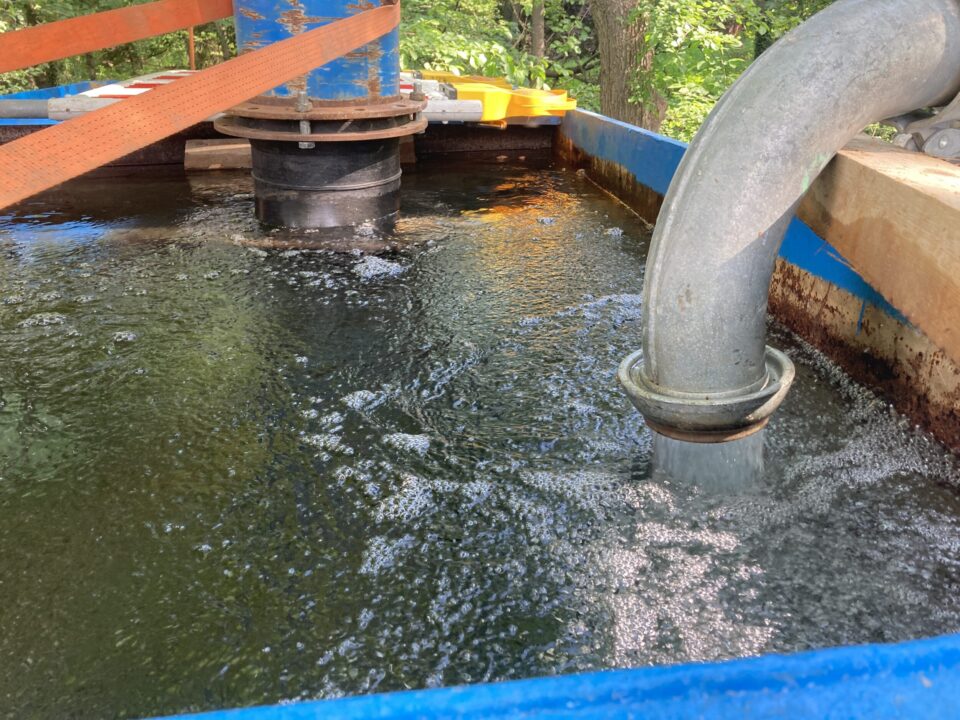 Das Bild zeigt einen großen blauen Wasserbehälter, der mit Wasser gefüllt ist und in dem sich deutliche Strömungen und Blasen an der Oberfläche bilden. Rechts mündet ein dickes Metallrohr in den Behälter und leitet Wasser hinein, während im Hintergrund ein weiteres Rohr und orangefarbene Gurte zur Befestigung sichtbar sind. Die Szene befindet sich im Freien, umgeben von grünem Laub und Bäumen.