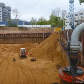 Das Bild zeigt eine große Baugrube mit sandigem Boden, die von einer hohen Spundwand aus Stahl umgeben ist. Im Vordergrund ist ein dickes Rohr mit einer roten Basis an der Wand befestigt, das vermutlich Teil einer Wasserhaltungseinrichtung ist. In der Grube stehen zwei Bagger, einer nahe der Wand und ein kleinerer auf einem Sandhügel, während im Hintergrund ein mehrstöckiges Gebäude, Container und ein Baukran zu sehen sind.