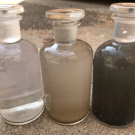 Das Bild zeigt drei Glasflaschen mit Glasstopfen, die nebeneinander auf einem Betonboden stehen. Jede Flasche enthält Wasserproben mit unterschiedlichen Trübungsgraden: links fast klar mit leichtem Bodensatz, in der Mitte milchig trüb und rechts deutlich dunkler mit starkem Sediment.