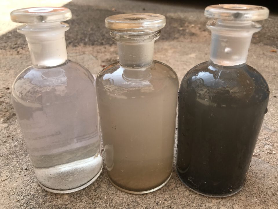 Das Bild zeigt drei Glasflaschen mit Glasstopfen, die nebeneinander auf einem Betonboden stehen. Jede Flasche enthält Wasserproben mit unterschiedlichen Trübungsgraden: links fast klar mit leichtem Bodensatz, in der Mitte milchig trüb und rechts deutlich dunkler mit starkem Sediment.