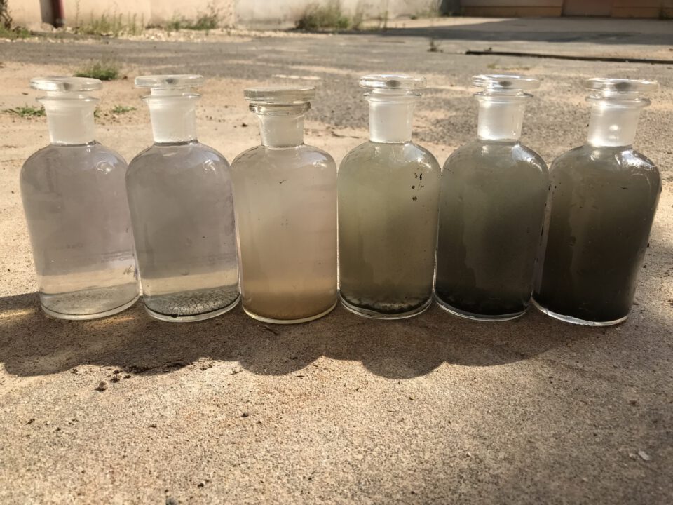 Das Bild zeigt sechs Glasflaschen mit breitem Hals und Glasstopfen, die nebeneinander auf einem sonnigen Betonboden stehen. Jede Flasche enthält Wasserproben mit unterschiedlichen Trübungsgraden: von fast klar links bis hin zu stark getrübt und dunkel rechts.