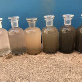 Das Bild zeigt sechs Glasflaschen mit Glasstopfen, die nebeneinander auf einer Betonfläche stehen. Jede Flasche enthält Wasserproben mit unterschiedlichen Trübungsgraden, von fast klar in der linken Flasche bis hin zu sehr dunklem, schlammigem Wasser in der rechten Flasche. Im Hintergrund ist eine blaue Fläche zu sehen, die einen einheitlichen Hintergrund für die Proben bildet.