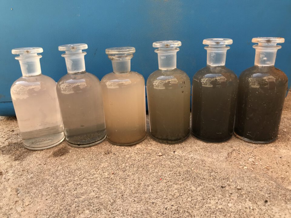 Das Bild zeigt sechs Glasflaschen mit Glasstopfen, die nebeneinander auf einer Betonfläche stehen. Jede Flasche enthält Wasserproben mit unterschiedlichen Trübungsgraden, von fast klar in der linken Flasche bis hin zu sehr dunklem, schlammigem Wasser in der rechten Flasche. Im Hintergrund ist eine blaue Fläche zu sehen, die einen einheitlichen Hintergrund für die Proben bildet.