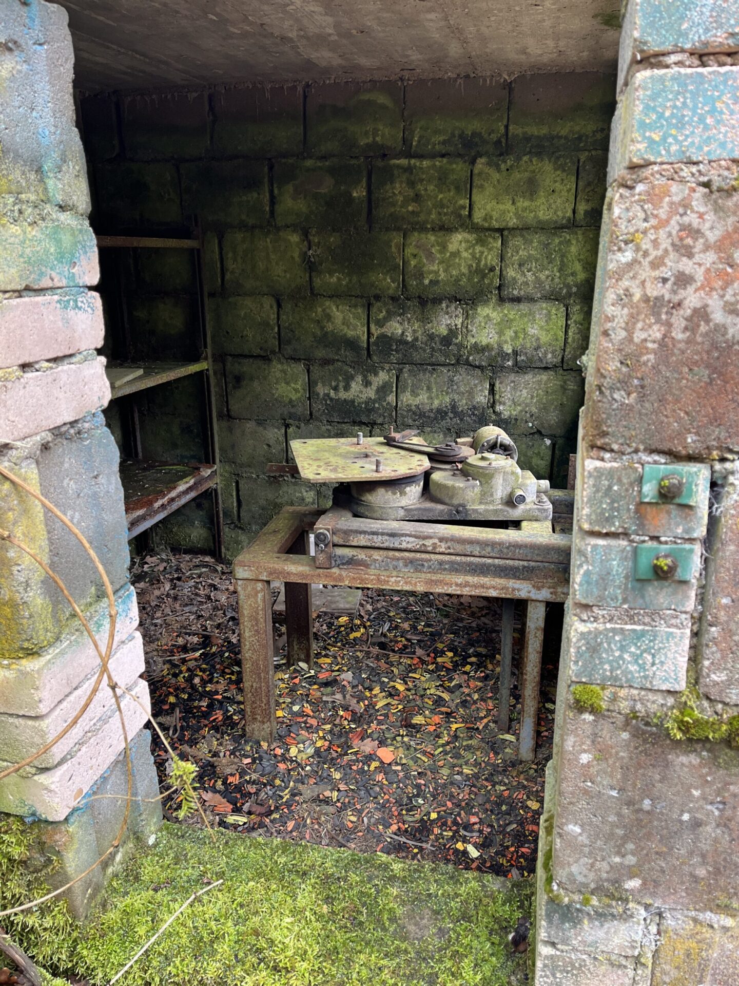 In einem offenen, verwitterten Unterstand steht eine stark verrostete Maschine mit mehreren runden Metallteilen und einer großen Platte. Die Umgebung wirkt verlassen, mit Moos an den Wänden und bunten Scherben am Boden.
