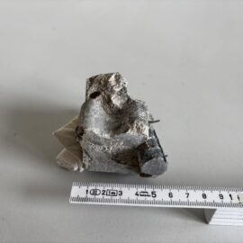 Das Bild zeigt ein unregelmäßig geformtes Stück Beton, das teilweise gebrochen ist und eine raue Oberfläche mit sichtbaren Bruchkanten aufweist. In dem Betonstück ist ein metallischer Abstandshalter oder Draht integriert, der an einer Seite herausragt. Unterhalb des Objekts liegt ein Maßband, das eine Länge von etwa 12 cm anzeigt, was die Größe des Bruchstücks verdeutlicht. Die Aufnahme erfolgt auf einer glatten, hellen Unterlage, die den Kontrast zum grauen Beton verstärkt.