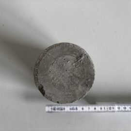 Das Bild zeigt einen zylindrischen Betonprobekörper, der von oben betrachtet wird. Die Oberfläche des Zylinders ist grau und weist eine raue, leicht unebene Struktur mit kleinen Poren und Abplatzungen auf. Unterhalb des Probekörpers liegt ein Maßband, das eine Länge von etwa 17 cm anzeigt, wobei die Skala auf die Größe des Zylinders hinweist. Die Aufnahme erfolgt auf einer hellen, glatten Unterlage, die den Kontrast zum Beton verstärkt.