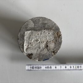 Das Bild zeigt einen zylindrischen Betonprobekörper, der von oben betrachtet wird. Die Oberfläche ist grau und weist eine unregelmäßige Struktur mit einem helleren, rechteckigen Bereich in der Mitte auf, der sich vom restlichen Material abhebt. Unterhalb des Probekörpers liegt ein Maßband, das eine Länge von etwa 14 cm anzeigt, was die Größe des Zylinders verdeutlicht. Die Aufnahme erfolgt auf einer glatten, hellen Unterlage, die den Kontrast zum Beton verstärkt.