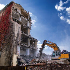 Ein gelber Bagger arbeitet am Abriss eines mehrstöckigen Gebäudes, dessen Fassade teilweise von roten Kletterpflanzen überwuchert ist. Der Bagger steht auf einem großen Schutthaufen aus Beton und Ziegeln, während Staub in der Luft liegt. Im Hintergrund ist ein blauer Himmel mit weißen Wolken zu sehen, und links im Bild befinden sich grüne Bäume.