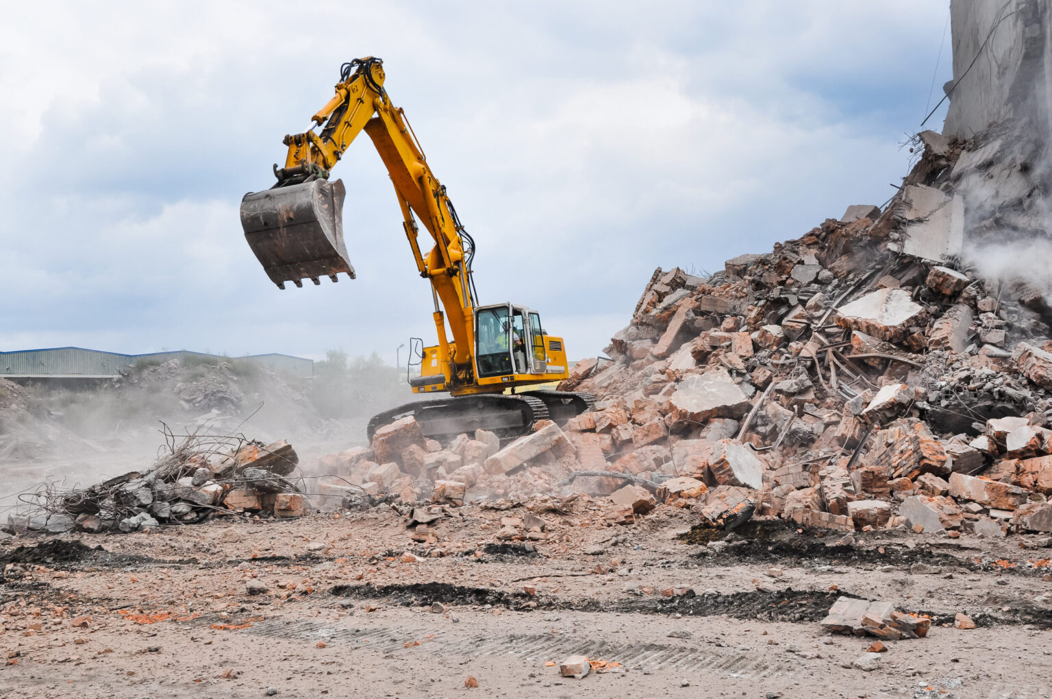 Ein gelber Bagger steht auf einem großen Schutthaufen aus zerbrochenem Beton und Ziegelsteinen. Der Bagger hebt seine Schaufel an, während Staub über den Trümmern liegt und Stahlarmierungen aus den Bruchstücken herausragen. Im Hintergrund ist ein wolkiger Himmel zu sehen, was die Szene offen und weit erscheinen lässt.