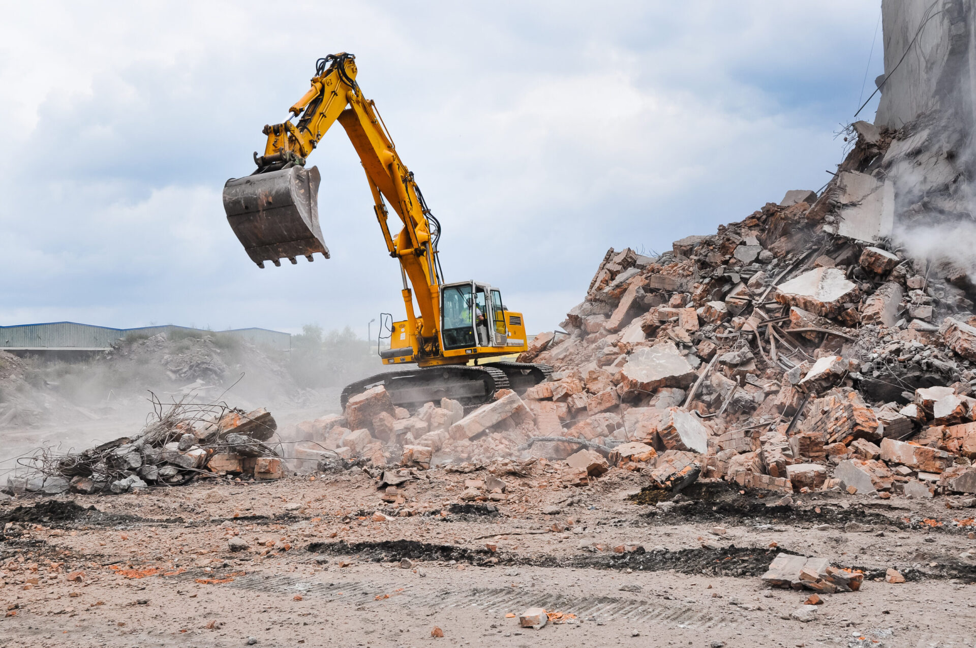 Ein gelber Bagger steht auf einem großen Schutthaufen aus zerbrochenem Beton und Ziegelsteinen. Der Bagger hebt seine Schaufel an, während Staub über den Trümmern liegt und Stahlarmierungen aus den Bruchstücken herausragen. Im Hintergrund ist ein wolkiger Himmel zu sehen, was die Szene offen und weit erscheinen lässt.