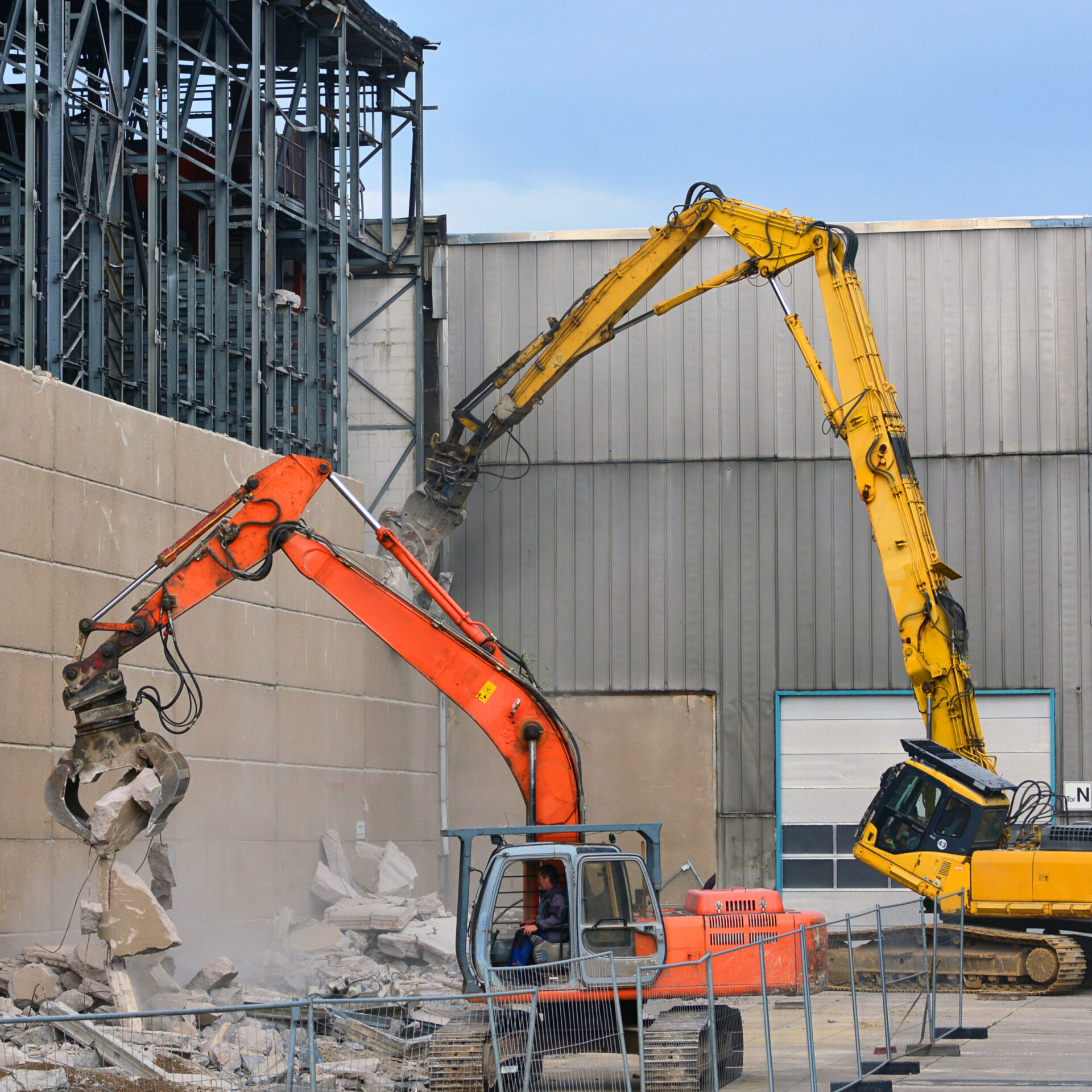Zwei große Bagger arbeiten an einer Abrissstelle vor einer hohen Betonwand. Der orangefarbene Bagger im Vordergrund zerkleinert die Wand, während der gelbe Bagger dahinter mit seinem langen Arm an einer höheren Stelle des Gebäudes ansetzt. Um die Maschinen liegen Betontrümmer, und im Hintergrund sind Metallkonstruktionen sowie eine große Industriehalle zu sehen.