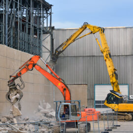 Zwei große Bagger arbeiten an einer Abrissstelle vor einer hohen Betonwand. Der orangefarbene Bagger im Vordergrund zerkleinert die Wand, während der gelbe Bagger dahinter mit seinem langen Arm an einer höheren Stelle des Gebäudes ansetzt. Um die Maschinen liegen Betontrümmer, und im Hintergrund sind Metallkonstruktionen sowie eine große Industriehalle zu sehen.