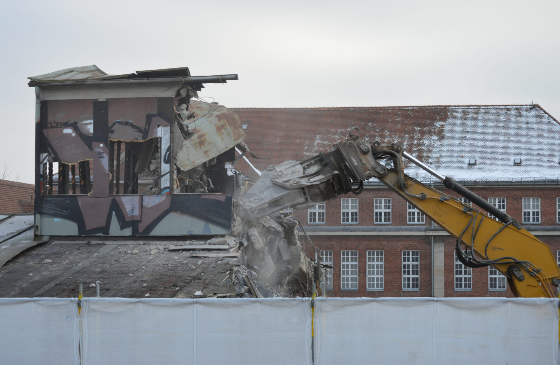Ein gelber Bagger reißt den oberen Teil eines Gebäudes ab, dessen Fassade mit großflächigen Graffiti bemalt ist. Der Greifarm des Baggers greift in die beschädigte Struktur, während Beton und Metallteile zerbrechen und Staub aufwirbelt. Im Hintergrund ist ein größeres Backsteingebäude mit vielen Fenstern und einem teilweise verschneiten Dach zu sehen.