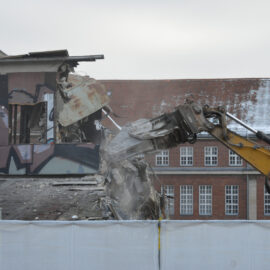 Ein gelber Bagger reißt den oberen Teil eines Gebäudes ab, dessen Fassade mit großflächigen Graffiti bemalt ist. Der Greifarm des Baggers greift in die beschädigte Struktur, während Beton und Metallteile zerbrechen und Staub aufwirbelt. Im Hintergrund ist ein größeres Backsteingebäude mit vielen Fenstern und einem teilweise verschneiten Dach zu sehen.
