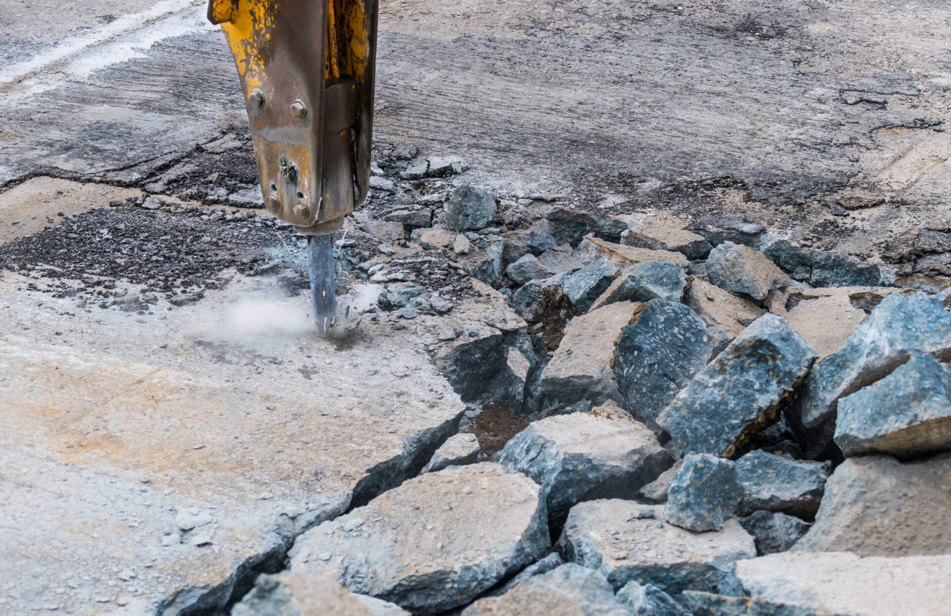 Ein hydraulischer Meißel bearbeitet eine Betonfläche und zerkleinert das Material in große, unregelmäßige Stücke. Rund um die Bruchstelle liegen bereits zahlreiche herausgebrochene Betonbrocken, während feiner Staub durch die Erschütterung entsteht. Die Oberfläche zeigt deutliche Risse und Bruchlinien, die den Fortschritt der Arbeiten sichtbar machen.