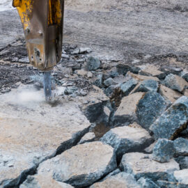 Ein hydraulischer Meißel bearbeitet eine Betonfläche und zerkleinert das Material in große, unregelmäßige Stücke. Rund um die Bruchstelle liegen bereits zahlreiche herausgebrochene Betonbrocken, während feiner Staub durch die Erschütterung entsteht. Die Oberfläche zeigt deutliche Risse und Bruchlinien, die den Fortschritt der Arbeiten sichtbar machen.