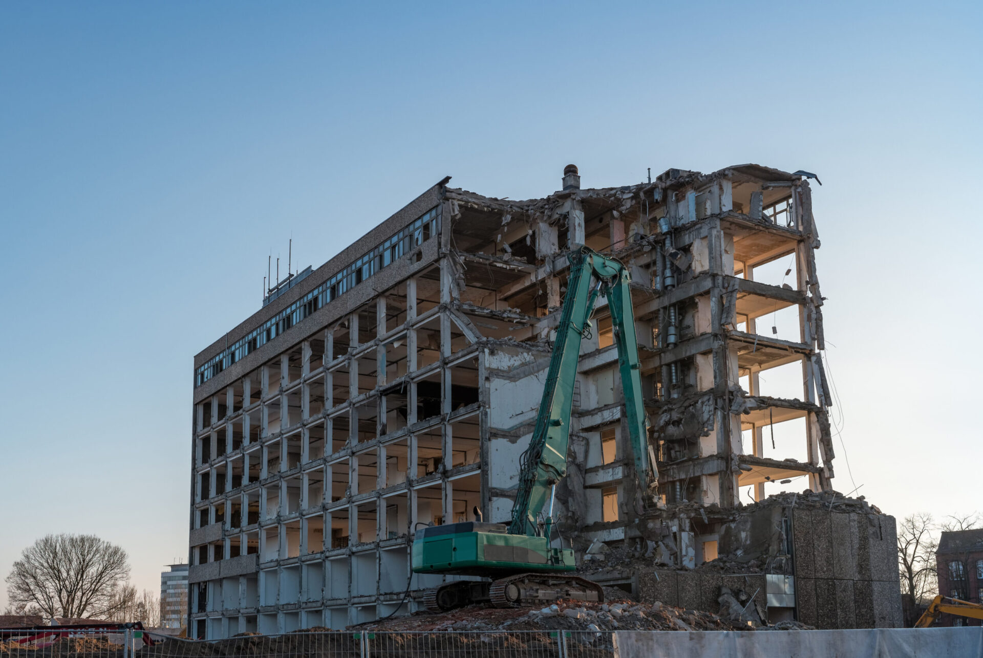 Ein großes mehrstöckiges Gebäude befindet sich im Abriss, wobei Teile der Fassade und Decken bereits entfernt sind. Vor dem Gebäude steht ein grüner Bagger mit langem Arm, der an der Struktur arbeitet und von Schutthaufen umgeben ist. Der Himmel ist klar und blau, und im Hintergrund sind einige Bäume und kleinere Gebäude zu erkennen.