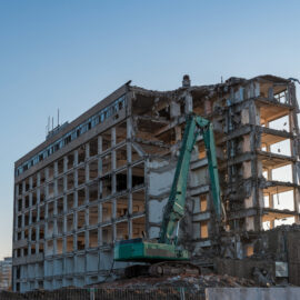 Ein großes mehrstöckiges Gebäude befindet sich im Abriss, wobei Teile der Fassade und Decken bereits entfernt sind. Vor dem Gebäude steht ein grüner Bagger mit langem Arm, der an der Struktur arbeitet und von Schutthaufen umgeben ist. Der Himmel ist klar und blau, und im Hintergrund sind einige Bäume und kleinere Gebäude zu erkennen.
