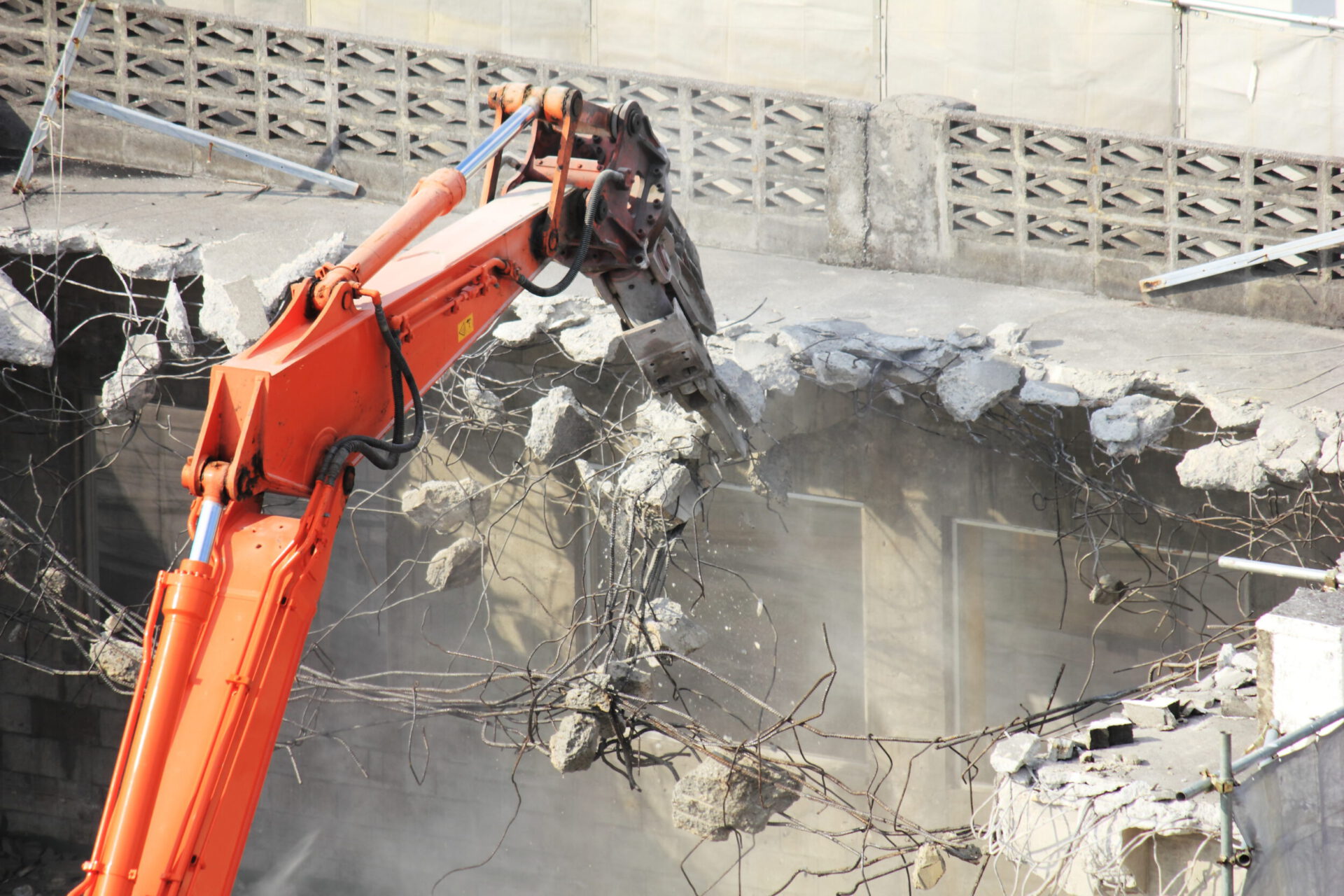 Ein großer orangefarbener Bagger ist dabei, eine massive Betonwand abzureißen. Der Greifarm des Baggers hält ein Stück der Wand, aus dem Stahlarmierungen herausragen. Um den Abrissbereich liegen zerbrochene Betonstücke und Staub, während im Hintergrund eine Mauer mit einem dekorativen Muster zu sehen ist.