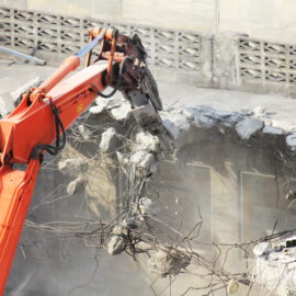 Ein großer orangefarbener Bagger ist dabei, eine massive Betonwand abzureißen. Der Greifarm des Baggers hält ein Stück der Wand, aus dem Stahlarmierungen herausragen. Um den Abrissbereich liegen zerbrochene Betonstücke und Staub, während im Hintergrund eine Mauer mit einem dekorativen Muster zu sehen ist.
