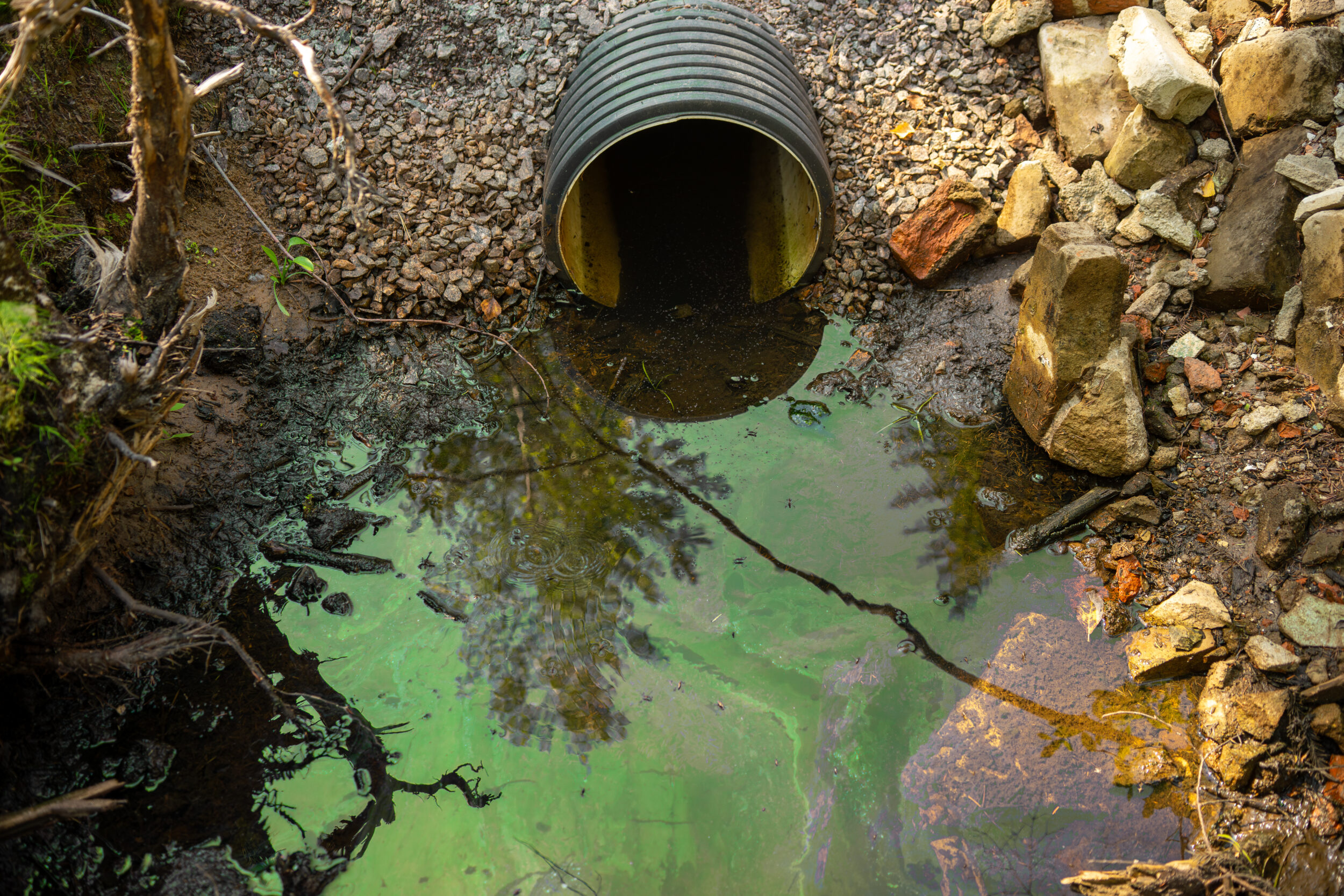 Das Bild zeigt einen Wasserlauf oder eine kleine Pfütze vor einem schwarzen Rohr, das aus einem steinigen Untergrund herausragt. Die Wasseroberfläche ist grünlich verfärbt und weist einen schimmernden Film auf, was auf eine mögliche Verschmutzung oder chemische Rückstände hindeutet. Rund um das Rohr liegen Steine und Erdmaterial, während im Wasser die Spiegelung von Ästen und Pflanzen sichtbar ist.