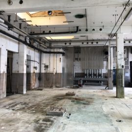 Das Bild zeigt einen leeren, stark abgenutzten Innenraum eines Industriegebäudes. Der Boden besteht aus alten, beschädigten Fliesen mit sichtbaren Spuren von Rost und Schmutz, während an den Wänden und der Decke zahlreiche Rohre und Kabelhalterungen verlaufen. Im Hintergrund sind technische Installationen und eine kleine Werkbank zu erkennen, was auf eine ehemalige Produktions- oder Maschinenhalle hinweist.