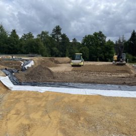 Das Bild zeigt eine große Baustelle mit vorbereiteten Erdflächen, die teilweise mit schwarzer Folie und weißem Vlies abgedeckt sind. In der Mitte befinden sich zwei Baumaschinen: eine gelbe Walze und ein Bagger, die auf einer Schicht aus Kies und Erde arbeiten. Im Hintergrund sind Bäume und ein bewölkter Himmel zu sehen, was auf eine offene Fläche in einem ländlichen Gebiet hinweist.