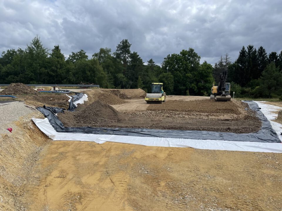 Das Bild zeigt eine große Baustelle mit vorbereiteten Erdflächen, die teilweise mit schwarzer Folie und weißem Vlies abgedeckt sind. In der Mitte befinden sich zwei Baumaschinen: eine gelbe Walze und ein Bagger, die auf einer Schicht aus Kies und Erde arbeiten. Im Hintergrund sind Bäume und ein bewölkter Himmel zu sehen, was auf eine offene Fläche in einem ländlichen Gebiet hinweist.