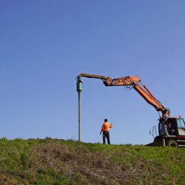 Das Bild zeigt eine große Baumaschine mit einem langen Ausleger, an dessen Ende ein senkrecht befestigter Bohrkopf angebracht ist. Daneben steht eine Person in auffälliger Arbeitskleidung auf einer grasbewachsenen Fläche und scheint die Arbeiten zu überwachen oder zu koordinieren. Der Hintergrund zeigt einen klaren blauen Himmel ohne weitere Strukturen, was die Szene offen und weitläufig wirken lässt.