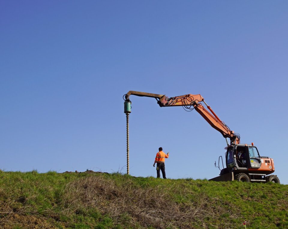 Das Bild zeigt eine große Baumaschine mit einem langen Ausleger, an dessen Ende ein senkrecht befestigter Bohrkopf angebracht ist. Daneben steht eine Person in auffälliger Arbeitskleidung auf einer grasbewachsenen Fläche und scheint die Arbeiten zu überwachen oder zu koordinieren. Der Hintergrund zeigt einen klaren blauen Himmel ohne weitere Strukturen, was die Szene offen und weitläufig wirken lässt.