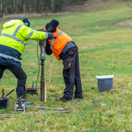 Das Bild zeigt zwei Personen in Arbeitskleidung, die auf einer Wiese mit einem technischen Gerät arbeiten. Sie tragen auffällige Sicherheitsjacken in Gelb und Orange und sind über ein metallisches Bohr- oder Messinstrument gebeugt, das senkrecht im Boden steht. Um sie herum liegen verschiedene Werkzeuge und Behälter, darunter ein weißer Kanister und eine graue Box, was auf eine Bodenuntersuchung oder Probebohrung im Freien hinweist.