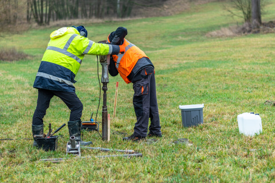 Das Bild zeigt zwei Personen in Arbeitskleidung, die auf einer Wiese mit einem technischen Gerät arbeiten. Sie tragen auffällige Sicherheitsjacken in Gelb und Orange und sind über ein metallisches Bohr- oder Messinstrument gebeugt, das senkrecht im Boden steht. Um sie herum liegen verschiedene Werkzeuge und Behälter, darunter ein weißer Kanister und eine graue Box, was auf eine Bodenuntersuchung oder Probebohrung im Freien hinweist.