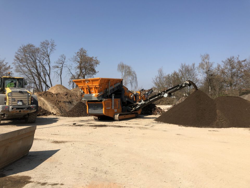 Das Bild zeigt eine große Siebanlage auf einer offenen Baustellenfläche, umgeben von mehreren Erdhaufen. Links ist ein Radlader zu sehen, der Material zur Weiterverarbeitung bereithält. Die Szene wirkt wie eine Anlage zur Aufbereitung oder Sortierung von Bodenmaterial im Freien.