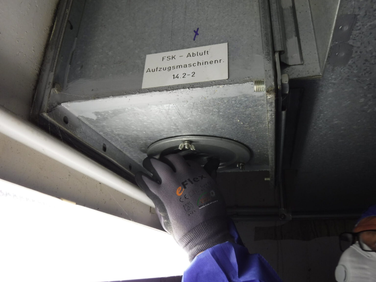 Das Bild zeigt eine Person, die mit einer Arbeitshandschuh bekleideten Hand an einem runden Metallteil arbeitet, das an einem großen Lüftungskanal oder einer Abluftanlage befestigt ist. Der Kanal besteht aus verzinktem Metall und ist mit einer Beschriftung versehen, die auf die Abluft einer Aufzugsmaschine hinweist. Im Vordergrund ist eine helle Leuchtstoffröhre zu sehen, die den Arbeitsbereich ausleuchtet, und die Person trägt blaue Schutzkleidung sowie Schutzausrüstung.