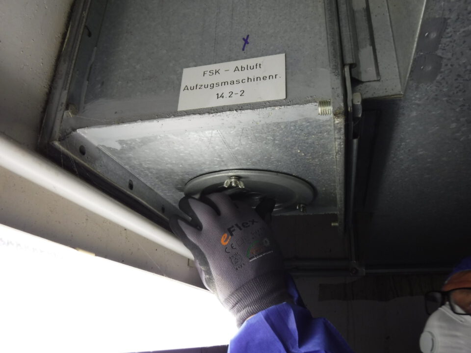Das Bild zeigt eine Person, die mit einer Arbeitshandschuh bekleideten Hand an einem runden Metallteil arbeitet, das an einem großen Lüftungskanal oder einer Abluftanlage befestigt ist. Der Kanal besteht aus verzinktem Metall und ist mit einer Beschriftung versehen, die auf die Abluft einer Aufzugsmaschine hinweist. Im Vordergrund ist eine helle Leuchtstoffröhre zu sehen, die den Arbeitsbereich ausleuchtet, und die Person trägt blaue Schutzkleidung sowie Schutzausrüstung.