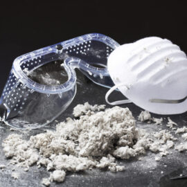 Das Bild zeigt eine weiße Atemschutzmaske und eine transparente Schutzbrille, die auf einer dunklen Oberfläche liegen. Davor befindet sich ein Haufen grauer, faseriger Materialien, die wie Baustoffreste oder Isoliermaterial wirken. Die Szene vermittelt den Eindruck von Arbeitsschutzmaßnahmen bei Tätigkeiten mit potenziell gefährlichen oder staubigen Materialien.