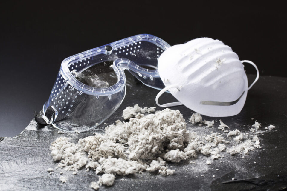 Das Bild zeigt eine weiße Atemschutzmaske und eine transparente Schutzbrille, die auf einer dunklen Oberfläche liegen. Davor befindet sich ein Haufen grauer, faseriger Materialien, die wie Baustoffreste oder Isoliermaterial wirken. Die Szene vermittelt den Eindruck von Arbeitsschutzmaßnahmen bei Tätigkeiten mit potenziell gefährlichen oder staubigen Materialien.