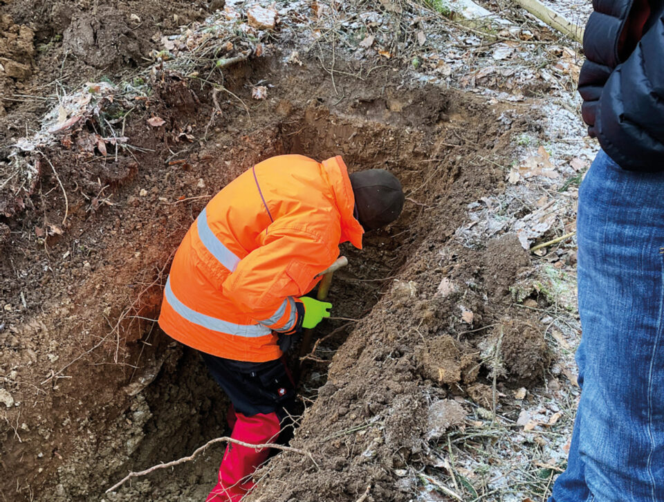 Das Bild zeigt eine Person in auffälliger Schutzkleidung, die in einer schmalen Erdgrube mit einem Werkzeug arbeitet. Der Boden ist aufgebrochen, Wurzeln und Erdschichten sind deutlich sichtbar. Daneben steht eine weitere Person am Rand der Grube und beobachtet die Arbeiten.