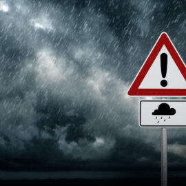 Das Bild zeigt ein dunkles, wolkenverhangenes Unwetter mit starkem Regen, der diagonal über die Szene fällt. Im Vordergrund steht ein Verkehrszeichen mit einem roten Dreieck und einem schwarzen Ausrufezeichen, darunter ein rechteckiges Schild mit einer stilisierten Regenwolke und Regentropfen. Die Kombination aus bedrohlichem Himmel und Warnschild vermittelt den Hinweis auf gefährliche Wetterbedingungen wie Starkregen oder Hochwasser.