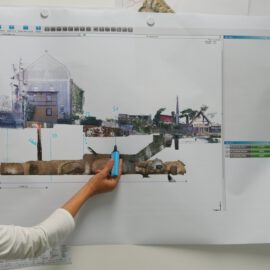 Das Bild zeigt eine große, ausgedruckte technische Darstellung an einer Wand, auf der ein Querschnitt eines unterirdischen Bauwerks mit darüberliegenden Gebäuden zu sehen ist. Eine Person hält mit der Hand einen Stift an eine markierte Stelle der Darstellung. Die Szene wirkt wie eine Besprechung oder Analyse eines Bau- oder Leitungsplans.