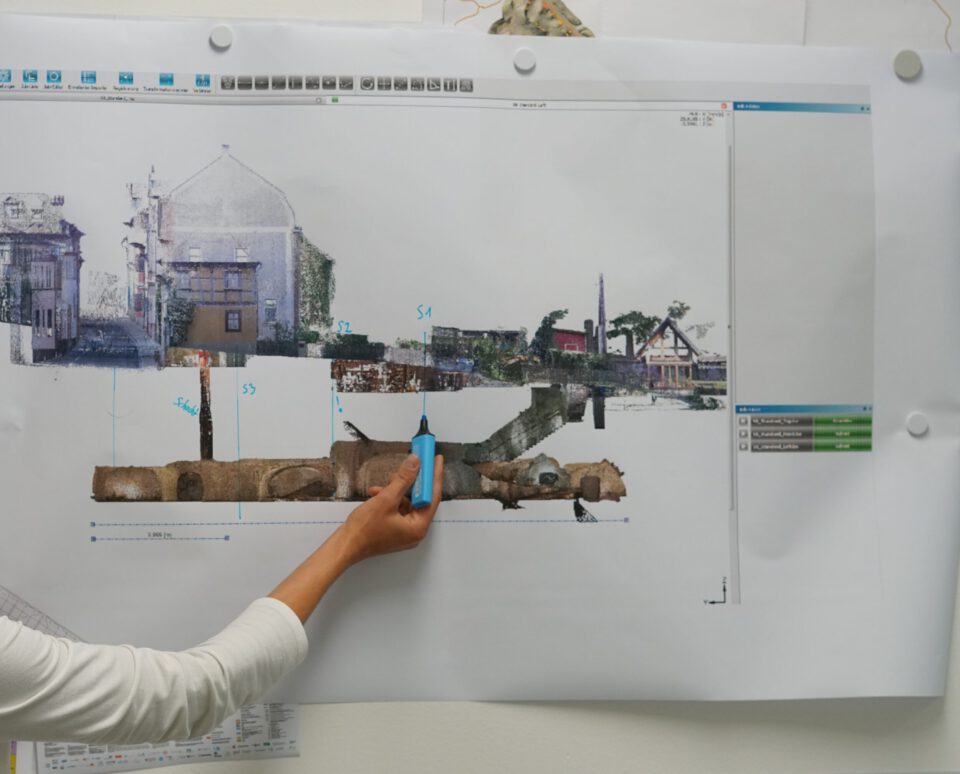 Das Bild zeigt eine große, ausgedruckte technische Darstellung an einer Wand, auf der ein Querschnitt eines unterirdischen Bauwerks mit darüberliegenden Gebäuden zu sehen ist. Eine Person hält mit der Hand einen Stift an eine markierte Stelle der Darstellung. Die Szene wirkt wie eine Besprechung oder Analyse eines Bau- oder Leitungsplans.