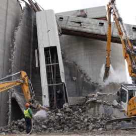 Das Bild zeigt den Abriss eines großen Betonbauwerks mit zwei schweren Baggern, die Trümmer abtragen. Ein Arbeiter in Schutzkleidung steht vor dem Schutt und hält einen Schlauch, während Staub aufwirbelt. Die eingestürzten Betonwände und freiliegenden Stahlstreben verdeutlichen den fortgeschrittenen Zustand der Abbrucharbeiten.
